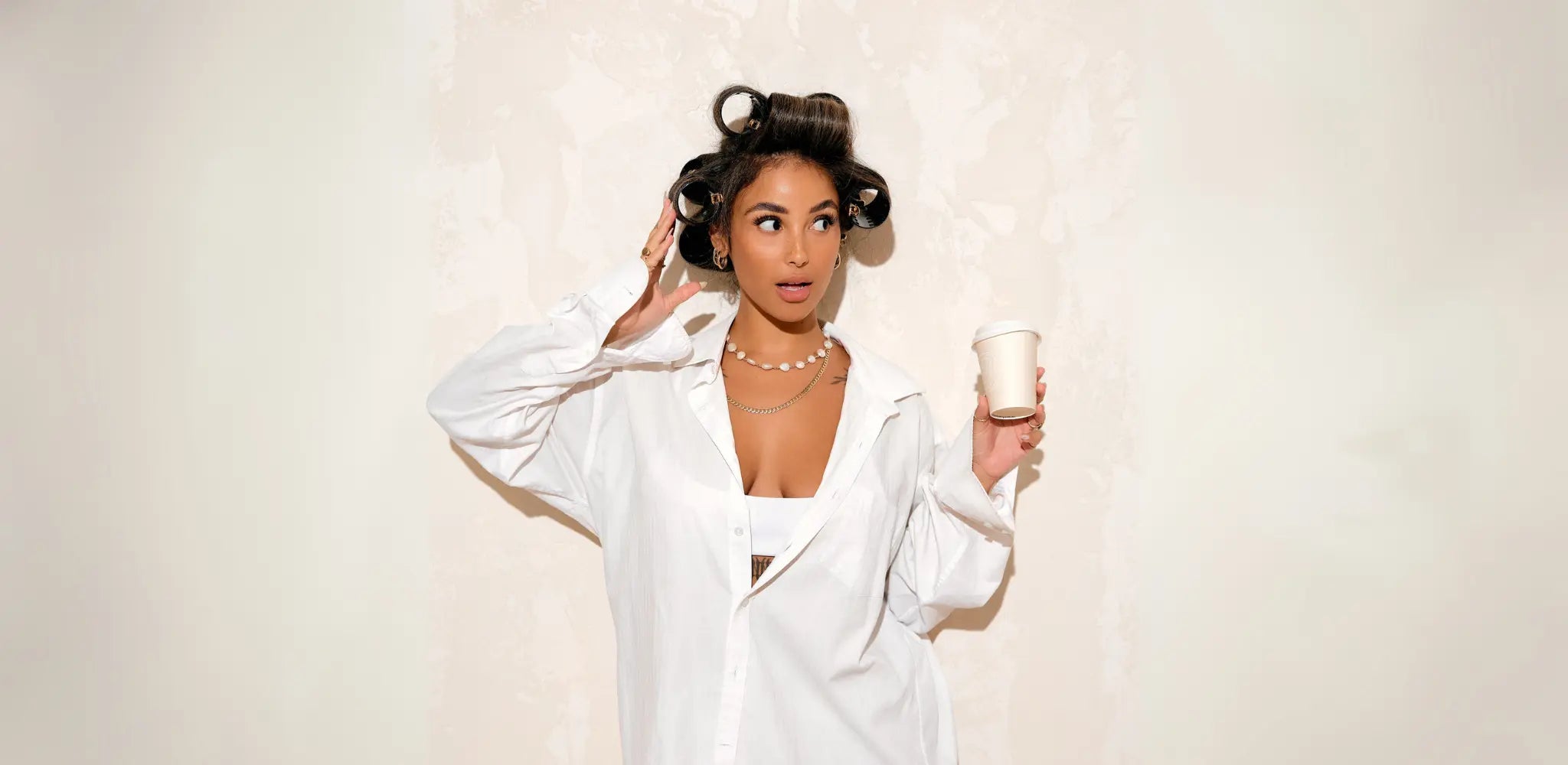 Woman with a surprised expression, her curly brown hair set in large Foxy Blondes hair rollers, wearing a white shirt and holding a coffee cup, looking off to the side.