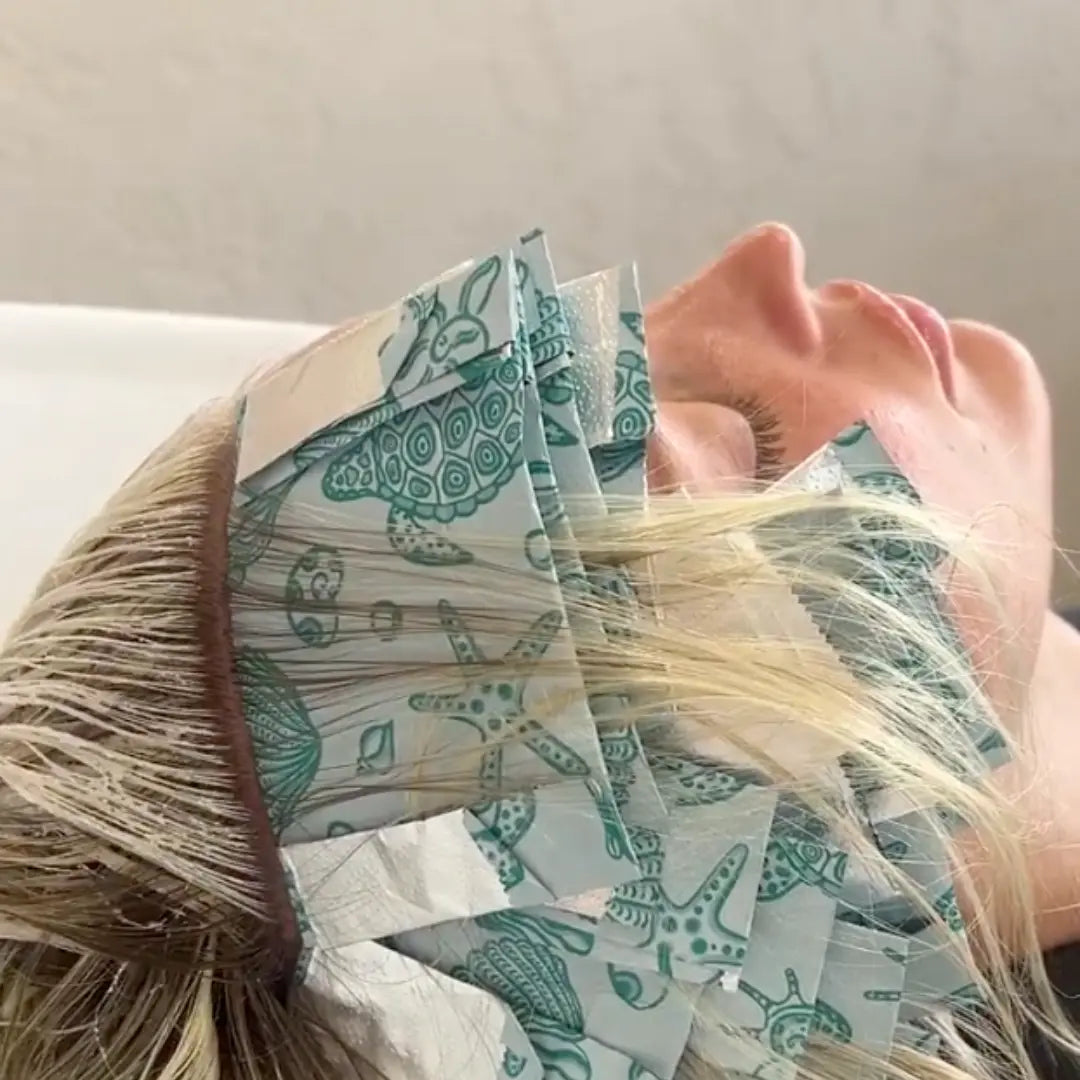 Close-up of Foxy Blondes 'Ocean Crush' patterned hair foils with turtle, starfish, and seashell designs applied to blonde hair, processing at a salon washbasin during a highlighting treatment.