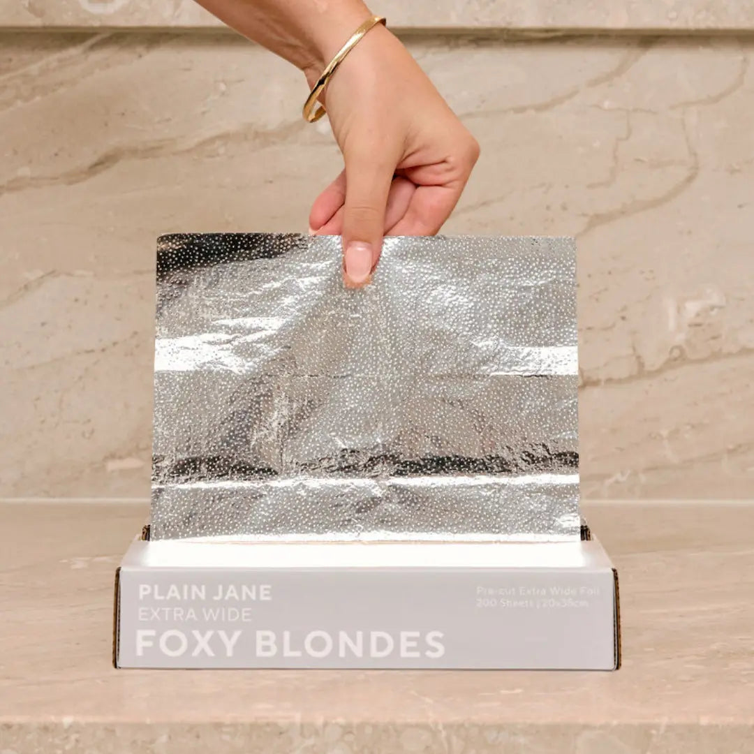 Hand pulling a Foxy Blondes 'Plain Jane' Extra Wide silver embossed pre-cut hair foil (20x35cm) from its 200-sheet dispenser box on a marble surface.