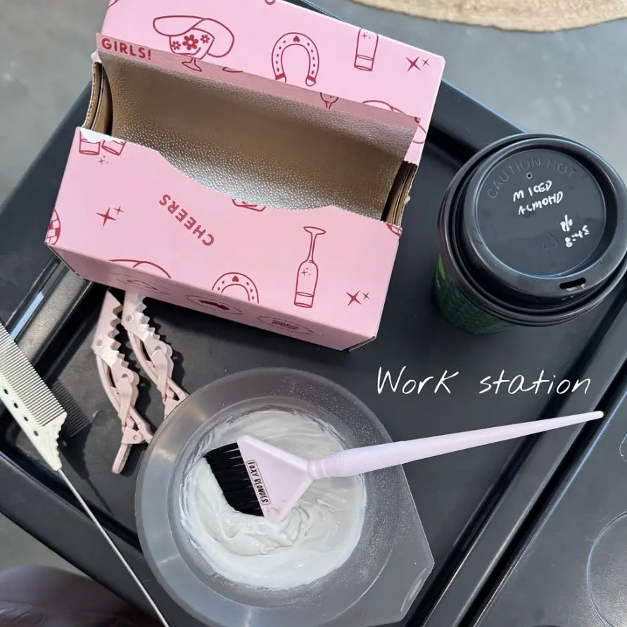 Hairstylist's salon workstation featuring an open box of Foxy Blondes 'Saddle Up' pink patterned foils, a Foxy Blondes tint brush in a bowl of hair colour, clips, comb, and a coffee cup on a trolley.